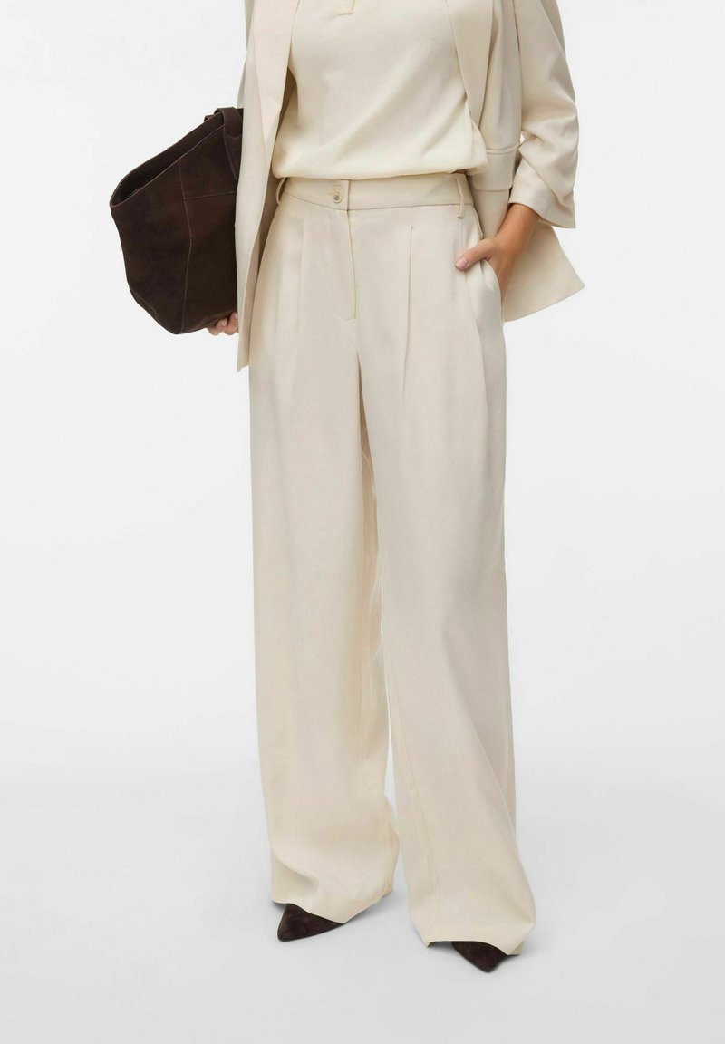 Pantalon large beige sur mesure avec plis, assorti à un top coordonné et un sac en daim marron foncé. Tissu lisse, design minimaliste.