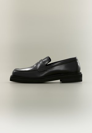 Čierny kožený penny loafer s okrúhlou špičkou, prešitými detailmi a nízkym vrstveným podpätkom na jednoduchom neutrálnom pozadí.