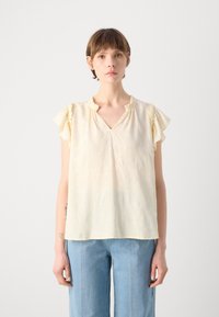Gul loftegenskjorte med V-hals og korte volangermer. Materialet har et subtilt mønster. Kombinert med lyseblå vide jeans.