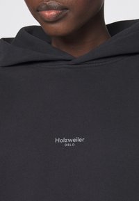 Gros plan d'une personne à la peau foncée portant un sweat à capuche noir avec « Holzweiler OSLO » écrit en petites lettres blanches sur la poitrine.