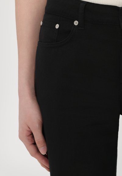 Sorte denim jeans med slim fit, med sølvfargede knapper, frontlommer og en strukturert overflate. Hånd som hviler mot jeansene.
