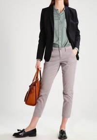 Kobieta w czarnym żakiecie, zielonej koszuli na guziki, jasnoszarych spodniach 3/4, czarnych mokasynach, trzymająca brązową skórzaną torebkę.