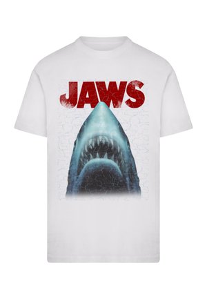T-shirt bianco con un grafico della bocca aperta di uno squalo e il testo "JAWS" in rosso in grassetto. Il design include una texture screpolata sullo sfondo.