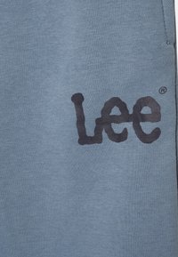 La tela de algodón azul presenta un prominente logo negro de "Lee". La textura es suave y uniforme, sin patrones adicionales ni acentos.