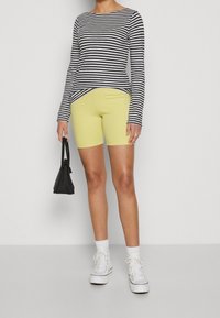 Camicia a maniche lunghe a righe in blu navy e bianco, abbinata a pantaloni corti gialli da ciclismo. Una borsa nera e scarpe da ginnastica bianche completano l'outfit.