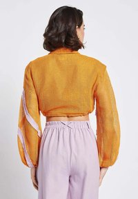 Giacca corta in rete arancione con maniche lunghe e dettagli a righe bianche. Abbinata a pantaloni elasticizzati viola chiaro per un fit rilassato.