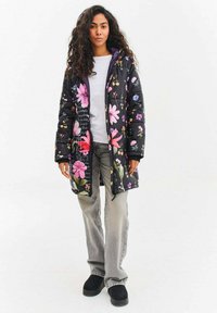 Květovaná prošívaná bunda s černým pozadím, na které jsou barevné květy, zapínání na zip vpředu a kapuce, doplněná o šedé kalhoty a černé boty.