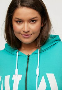 Sudadera con cremallera en color turquesa con un estampado gráfico en blanco, que incluye una capucha con cordón y una textura suave. Primer plano de la cremallera y los detalles.