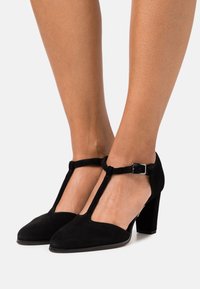 Chaussures à talons hauts en daim noir avec un design à sangle T et une boucle carrée à la cheville, dotées d'un bout arrondi et d'une finition texturée.