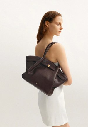 Femme en robe blanche portant deux grands sacs à main en cuir marron foncé sur son épaule, vue de profil sur un fond clair uni.