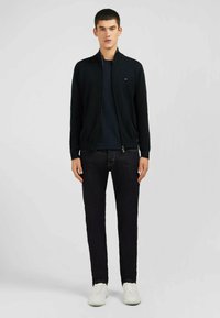 Veste noire zippée, avec des poignets côtelés, portée par-dessus une chemise sombre. Associée à un jean foncé et des baskets blanches. Texture lisse, design ajusté.