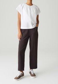 Personne portant un chemisier blanc à manches courtes, un pantalon ample foncé et des baskets marron, debout avec une main dans la poche devant un fond uni.