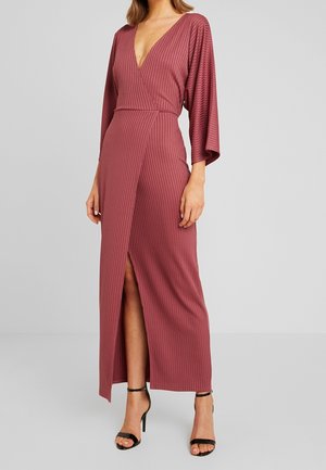 Abito lungo mauve a coste con scollatura a V profonda, maniche ampie e uno spacco alto fino alla coscia. Silhouette slanciata con vita curata da una cintura. Tacchi alti neri.