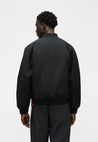 Άνθρωπος με κοντά σγουρά μαλλιά, φοράει μαύρο μπουφάν τύπου bomber και μαύρο παντελόνι, στέκεται με την πλάτη προς το μέρος μας μπροστά σε ένα απλό λευκό φόντο.