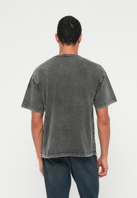Šedé nadměrně velké tričko z bavlny s opotřebovanou texturou, s krátkými rukávy a kontrastním prošitím podél lemů.