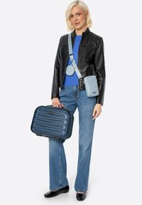 Valigia rigida blu scuro con design a righe verticali, abbinata a una borsa a tracolla blu chiaro e a una borsa rotonda, indossata da un modello.