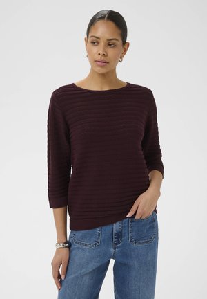 Vrouw met donker haar in een lage knot, draagt een donkerpaarse textuursweater met driekwartmouwen en blauwe spijkerbroek.