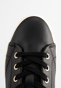 Baskets en cuir noir avec des lacets noirs passés dans des œillets dorés et une semelle en caoutchouc blanche visible à l'avant.