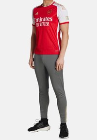 Red sport jersey with white sleeves, featuring a geometric pattern and "Emirates Fly Better" text. Paired with gray athletic pants and black shoes.