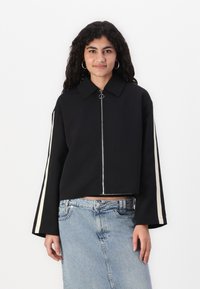 Veste noire courte avec un col pointu, fermeture éclair argentée et manches longues avec des rayures blanches sur les côtés. Texture lisse, design minimaliste.