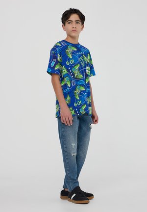 T-shirt grafica blu con design di controller di gioco verdi, abbinata a jeans in denim strappati e sneakers nere.
