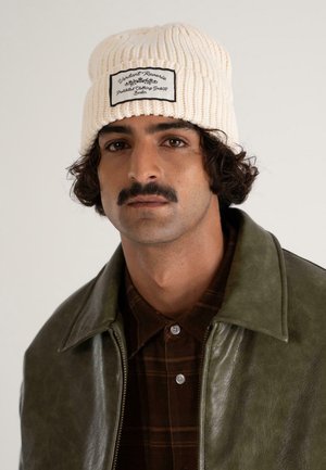 Uomo con capelli ricci e baffi che indossa un berretto di maglia crema con una toppa bianca e nera, giacca di pelle verde e camicia a quadri marrone.