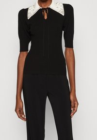 Haut noir côtelé avec un col blanc orné, des manches courtes bouffantes et un fermoir à nouer au niveau du cou. Présente une coupe ajustée et une texture lisse.