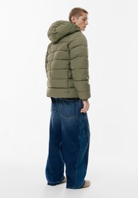 Giubbino imbottito verde oliva con cappuccio, caratterizzato da un design trapuntato. Indossato sopra dei jeans in denim blu scuro, larghi.