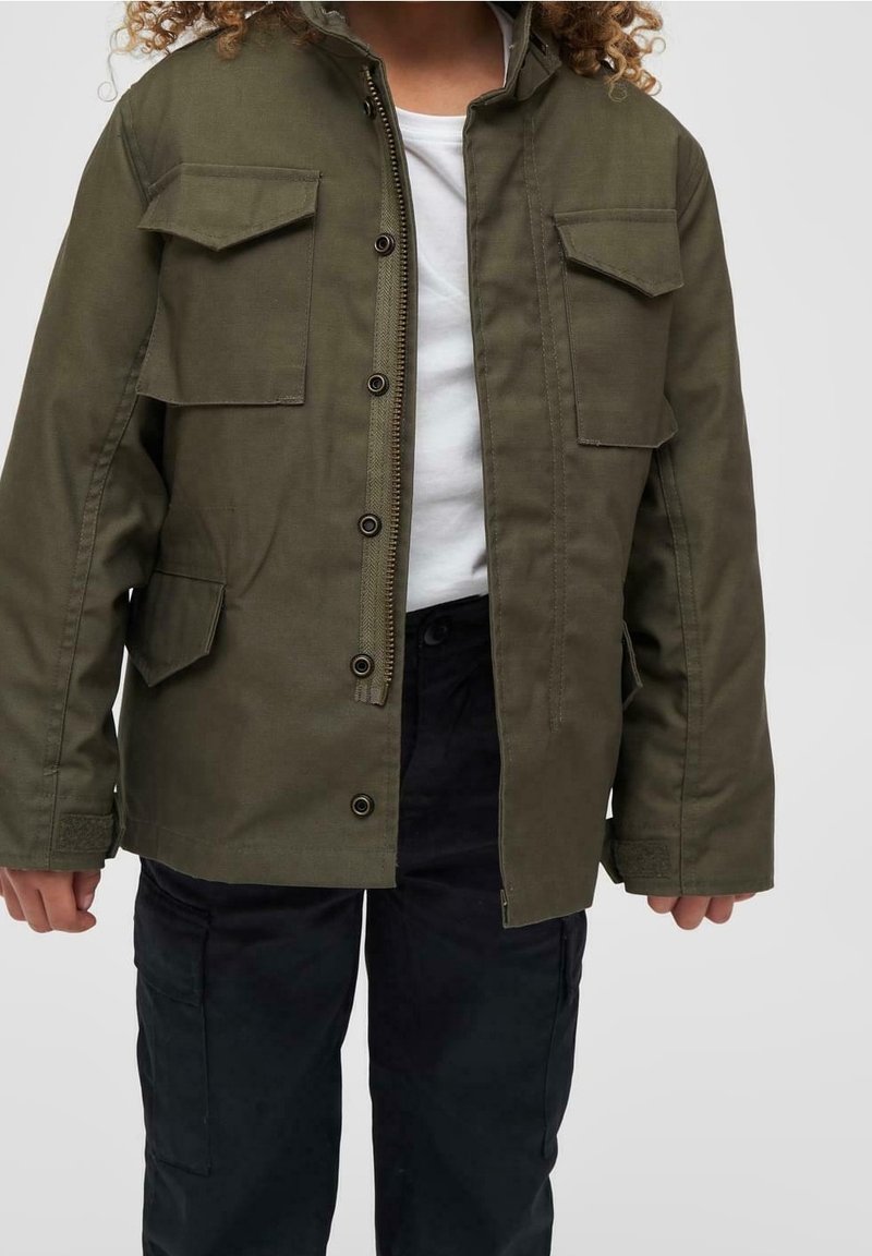 Enfant portant une veste vert olive ouverte sur une chemise blanche et un pantalon noir, debout sur un fond gris uni.
