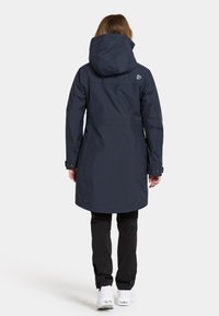 Lange marineblauwe waterdichte jas met een capuchon, voorzien van een trekkoord en een getailleerde taille. Gekoeld met zwarte broek en witte schoenen.