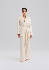 Slonovinovo satenové sako s prekladaným dizajnom, dlhými rukávmi a nohavicami so širokými nohavicami v rovnakom materiáli. Doplnené látkovým opaskom a jemnou textúrou.