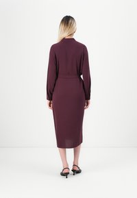 Woman with long blonde hair wearing a long-sleeve midi purple dress and black low-heeled shoes, standing with her back to the camera.
