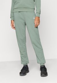 Jasnozielone spodnie dresowe o miękkiej fakturze, zwężanym kroju, elastycznych mankietach oraz małym białym logo na lewej biodrze, zestawione z czarnymi trampkami.
