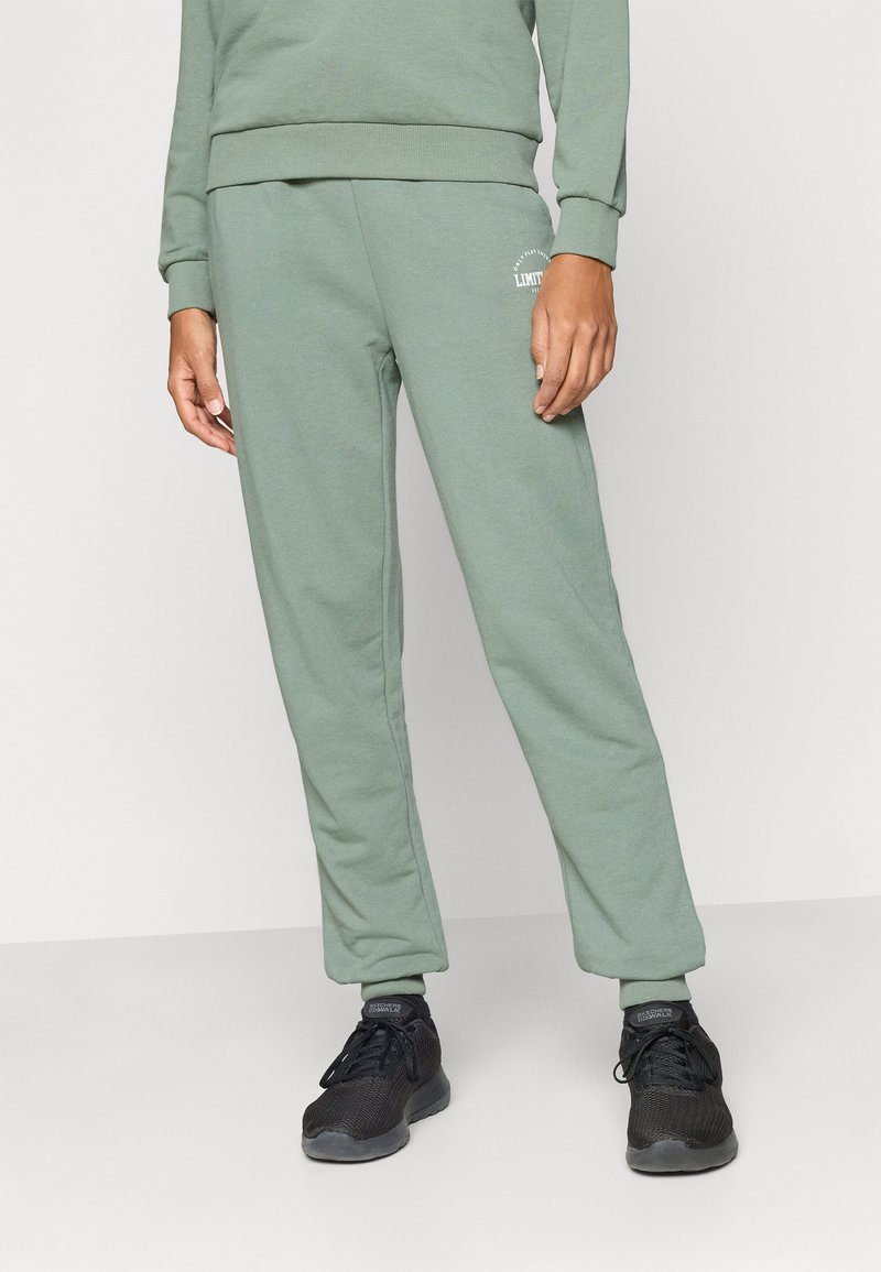 Jasnozielone spodnie dresowe o miękkiej fakturze, zwężanym kroju, elastycznych mankietach oraz małym białym logo na lewej biodrze, zestawione z czarnymi trampkami.