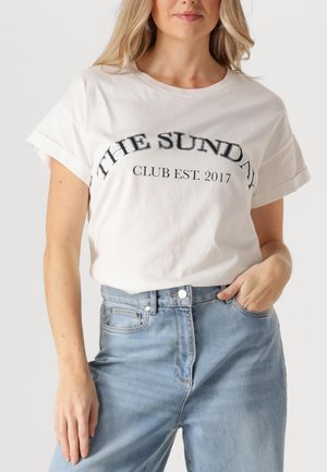 Witte katoenen t-shirt met opgerolde mouwen en gebogen tekst: "THE SUNDAY CLUB EST. 2017" in zwart en zilver gedrukt. Draag met lichtblauwe denim jeans.