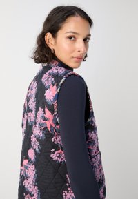 Gestepptes Weste mit einem floralen Muster in Pink und Lila auf einem dunklen Hintergrund. Die Ränder haben einen kontrastierenden Besatz.