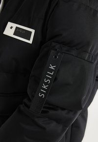 Svart vadderad jacka med en vit logotyp och en svart etikett med vit text på ärmen. Strukturerad tyg med en slät finish.