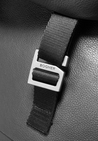 Črna teksturirana usnjena površina z črnim tkaninskim trakom in srebrno kovinsko zaponko z napisom "BOGNER". Povečani posnetek detajla strojne opreme.