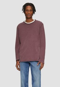 Langärmliges T-Shirt mit Rundhalsausschnitt in gedämpftem Mauve, mit strukturierter Oberfläche und sichtbaren Nähten. Kombiniert mit hellblauen, locker sitzenden Jeans.