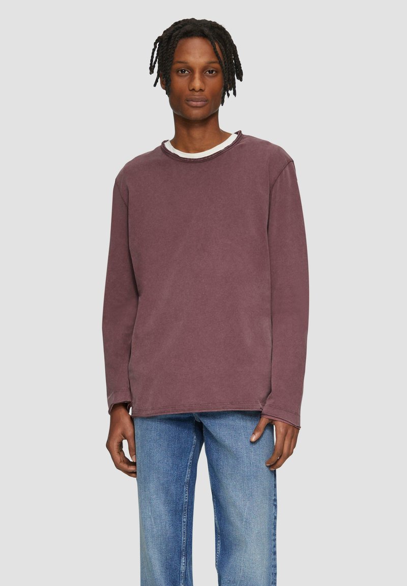 Langärmliges T-Shirt mit Rundhalsausschnitt in gedämpftem Mauve, mit strukturierter Oberfläche und sichtbaren Nähten. Kombiniert mit hellblauen, locker sitzenden Jeans.