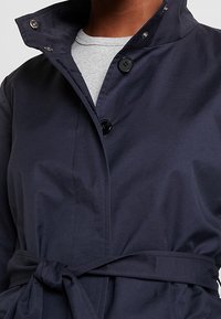 Cappotto trench blu navy con cintura e colletto alto, dotato di bottoni neri, texture liscia e design moderno.