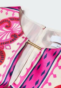 Blouse en soie avec un motif géométrique multicolore en rose, rouge et bleu, dotée d'un fermoir en barre dorée au niveau du col.