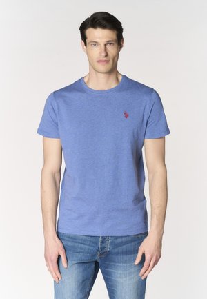 Uomo che indossa una semplice maglietta blu a maniche corte con un piccolo logo rosso sul petto, abbinata a jeans blu, in piedi davanti a uno sfondo bianco.