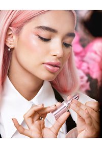 A person holds a clear, cylindrical lipstick tube with a metallic cap. The individual has pink hair and is wearing a white collared shirt.