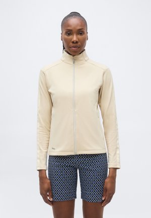 Woman with dark skin and hair tied back wears a beige zip-up jacket and blue patterned shorts, standing against a plain white background.