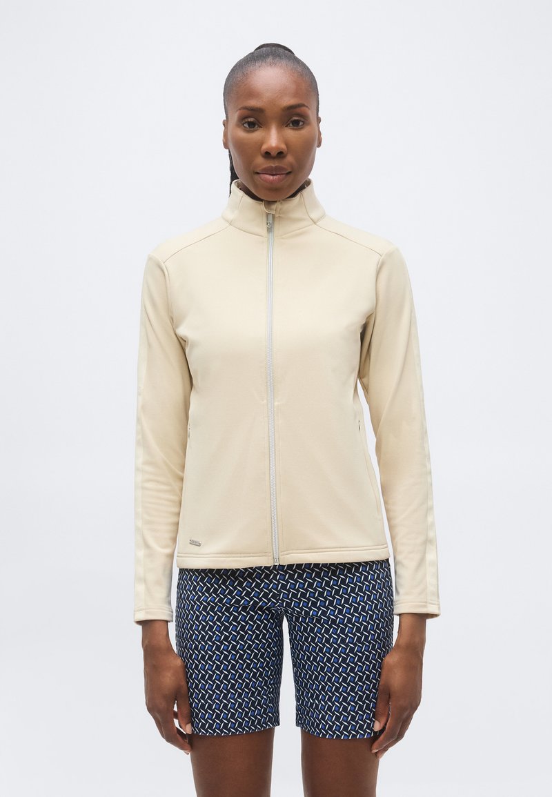 Γυναίκα με σκούρο δέρμα και μαλλιά δεμένα πίσω φοράει ένα μπεζ μπουφάν με φερμουάρ και μπλε σορτς με σχέδια, στέκεται μπροστά σε απλό λευκό φόντο.