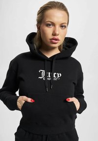 Černá mikina s kapucí vyrobená z měkké tkaniny, s přední kapsou, šňůrkami a logem "Lucy" v bílém písmu na hrudi.