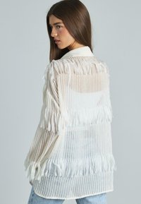 Weiße, transparente Bluse mit geraffter Textur, die horizontale Streifen und einen klassischen Kragen aufweist. Lange Ärmel reichen über die Handgelenke hinaus.