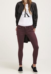Chaqueta ligera negra sobre una camiseta blanca con un logo negro, combinada con jeans ajustados burdeos y zapatillas negras sin cordones.
