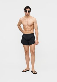 Särgil mees, kes kannab musta ujumispükst, musti päikeseprille ja musti plätusid, seisab ühevärvilise valge tausta ees.